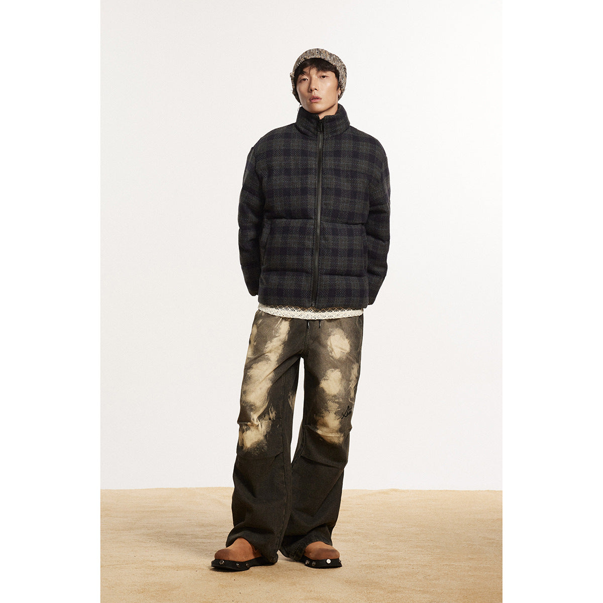 Front view of a model in a zipped dark plaid puffer jacket, cream knit layer, and mottled brown pants.