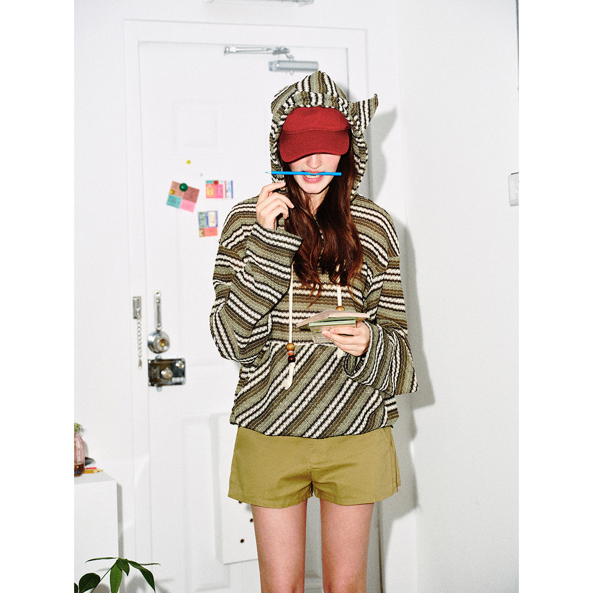 Model wearing an olive, cream and black striped hooded pullover with a red cap and olive shorts.