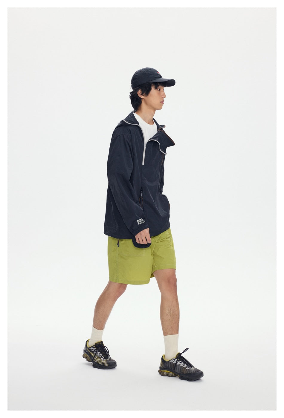Side view of model walking in black hooded jacket with diagonal zipper, green shorts and cap