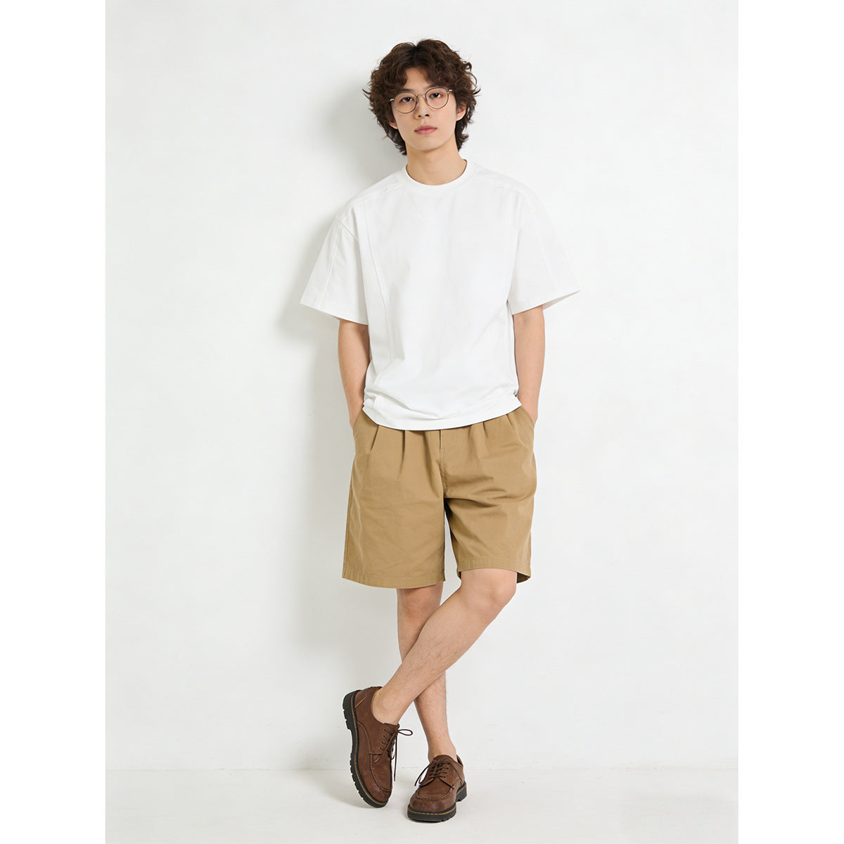 Model wearing a plain white short-sleeve T-shirt with tan shorts and brown lace-up shoes, full-length front view.