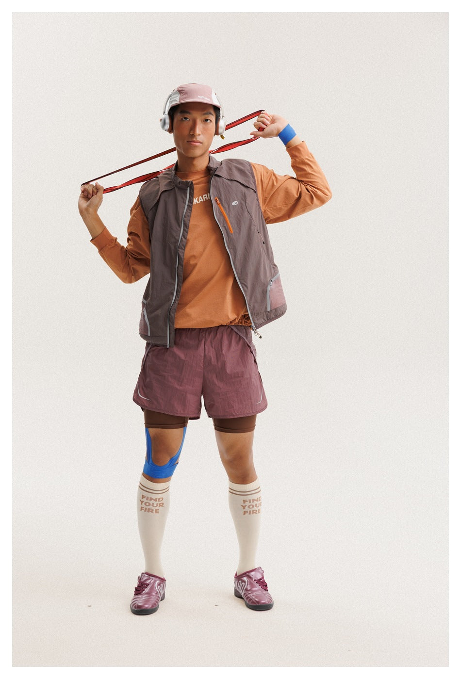 Full-body three-quarter view of a man wearing a taupe sleeveless vest over an orange top and mauve shorts.