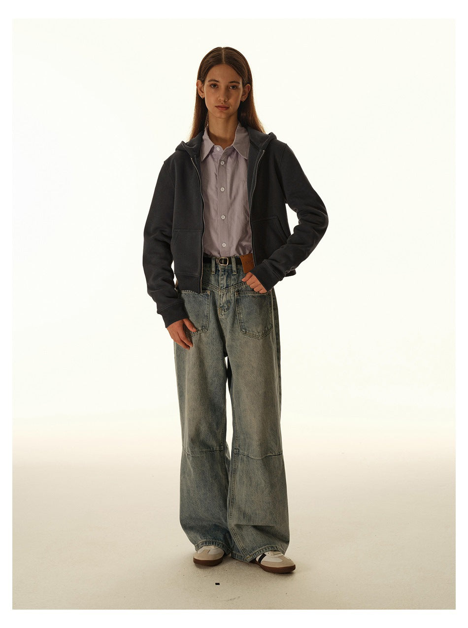 Model wearing a charcoal zip-front hooded jacket over a pale button-up shirt and loose gray-blue jeans.