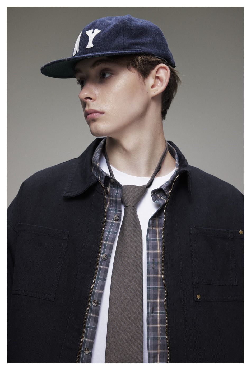Model wearing a dark work jacket over a plaid shirt and white tee, with a navy cap featuring a white NY emblem.