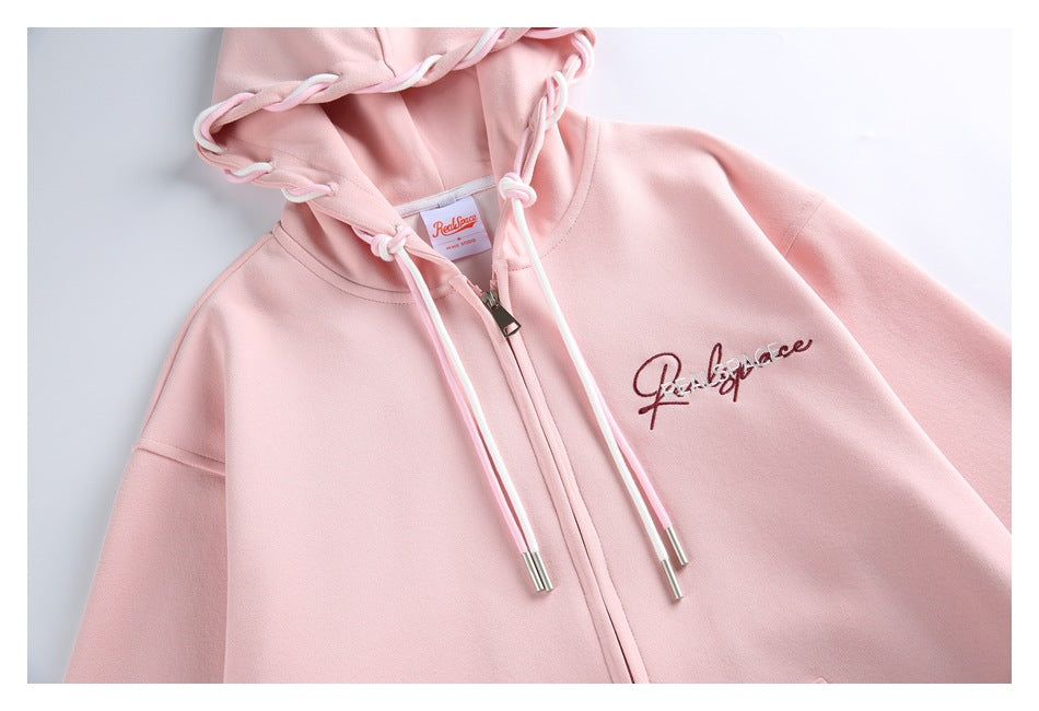 Flat lay of a pale pink zip-front hoodie with white drawstrings, a knotted hood edge and embroidered script logo.