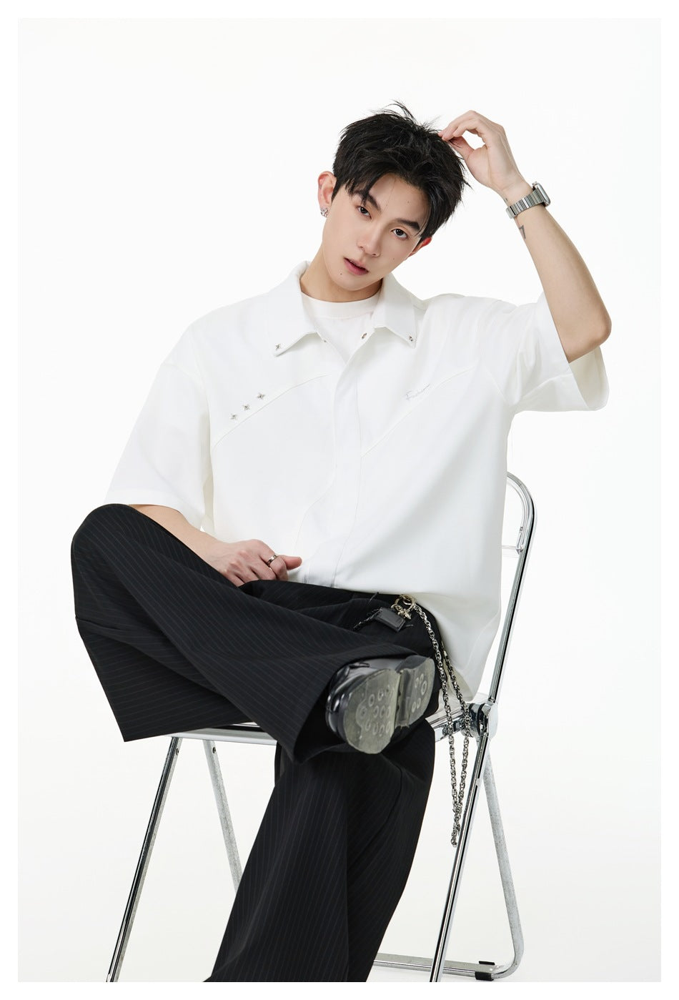 Seated model wearing an oversized white short-sleeve button-up shirt with curved seams and small embellishments.