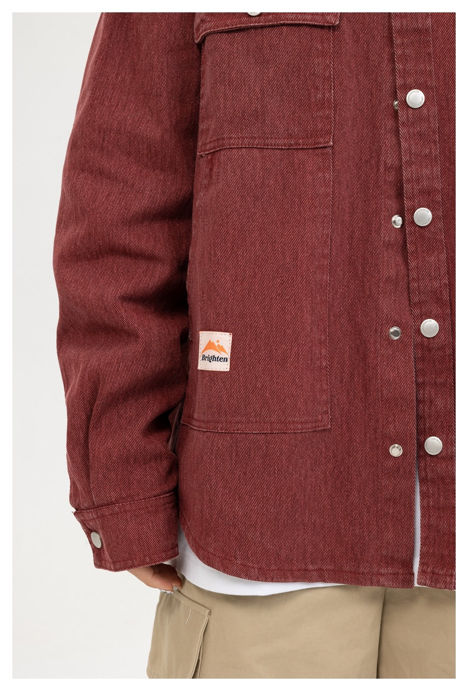 Close-up of a burgundy button-front jacket with a large patch pocket and small sewn label, worn over a white shirt.