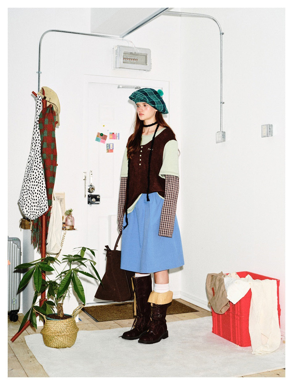 Side view of a model in a brown sleeveless top layered over shirts, a blue skirt, and a teal plaid beret.