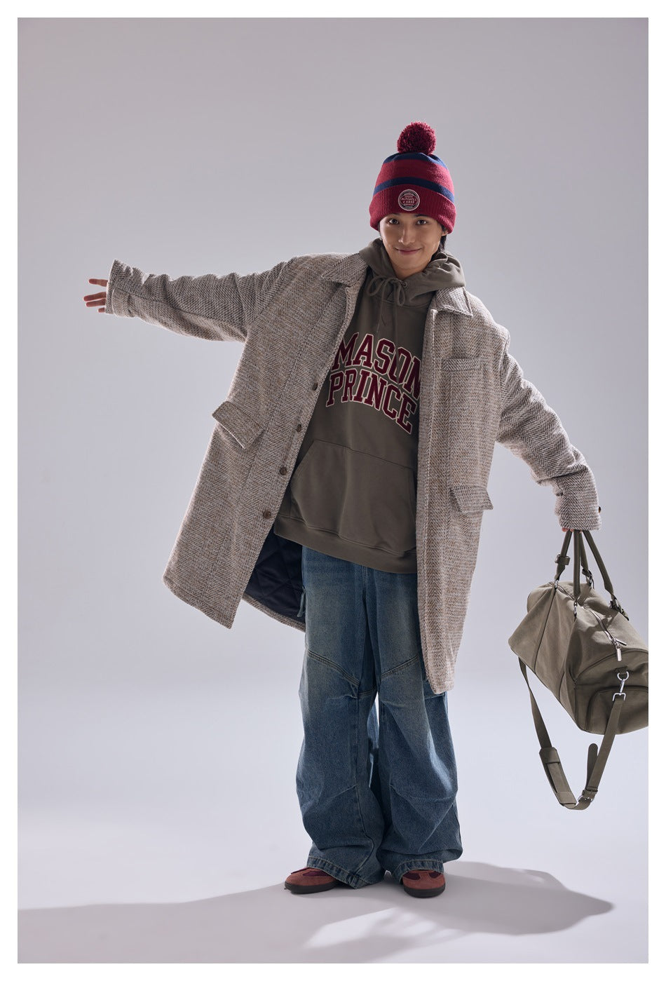 Model wearing a beige patterned long coat, printed hoodie, jeans, and red striped beanie, holding an olive duffel bag.
