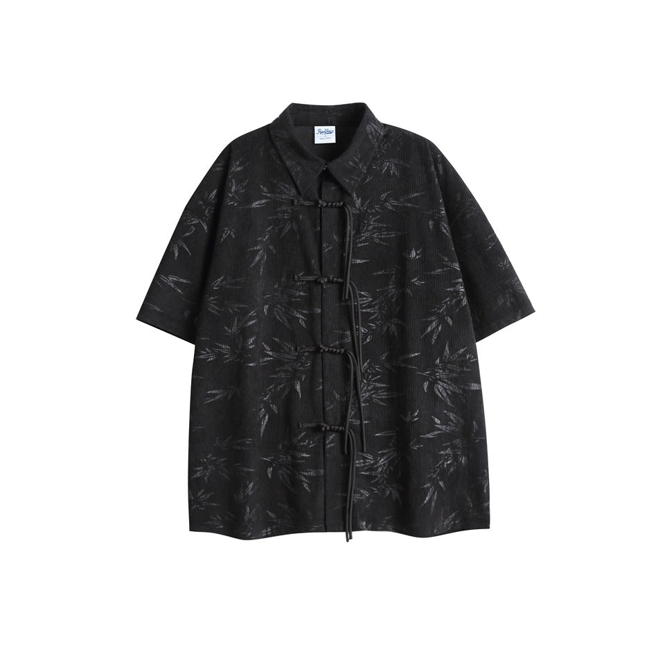 Flat-lay front view of a black short-sleeve collared tunic with a subtle leaf pattern and frog closures.