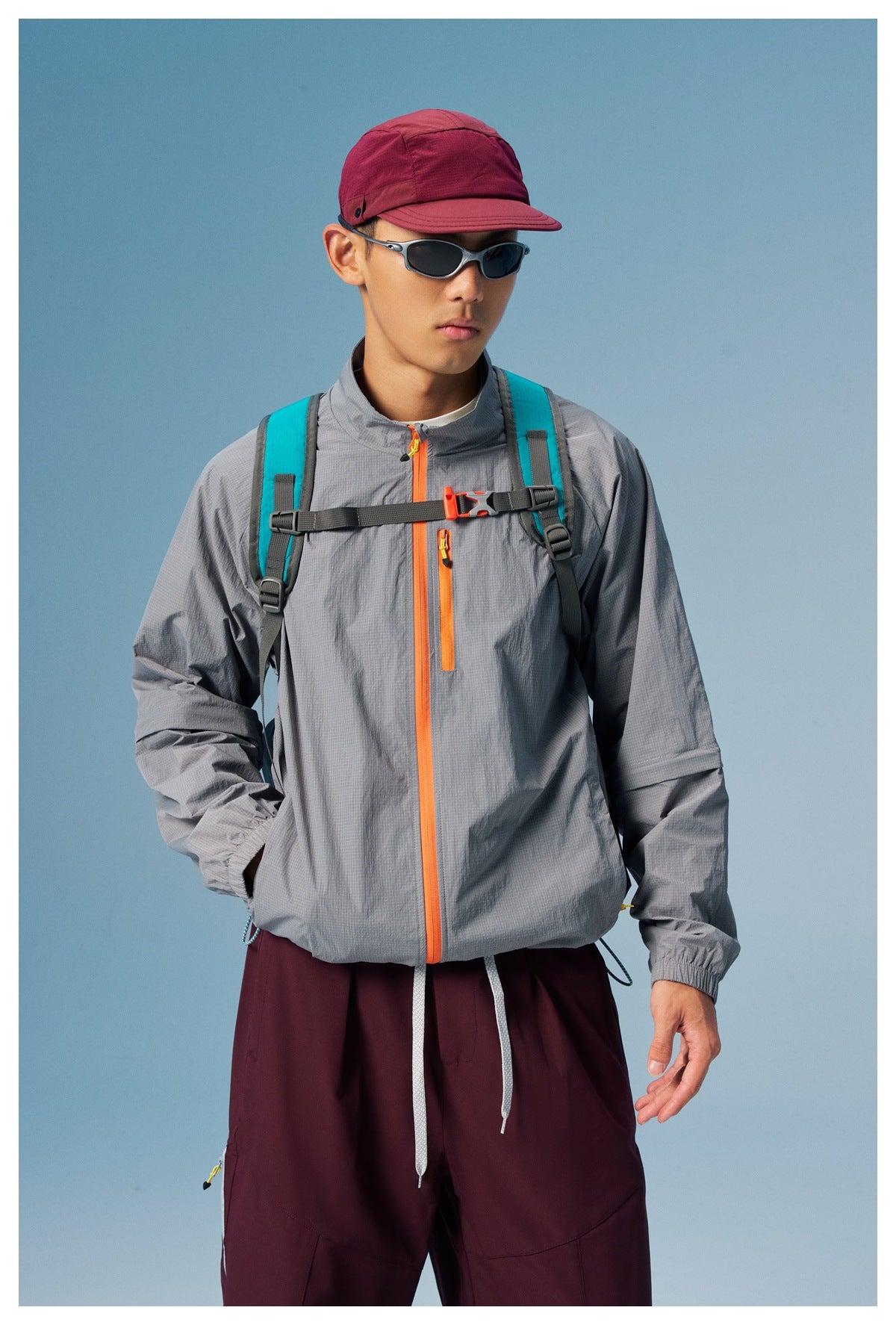 Closer front view of a model wearing a gray jacket with bright orange zips and a teal-strapped backpack.