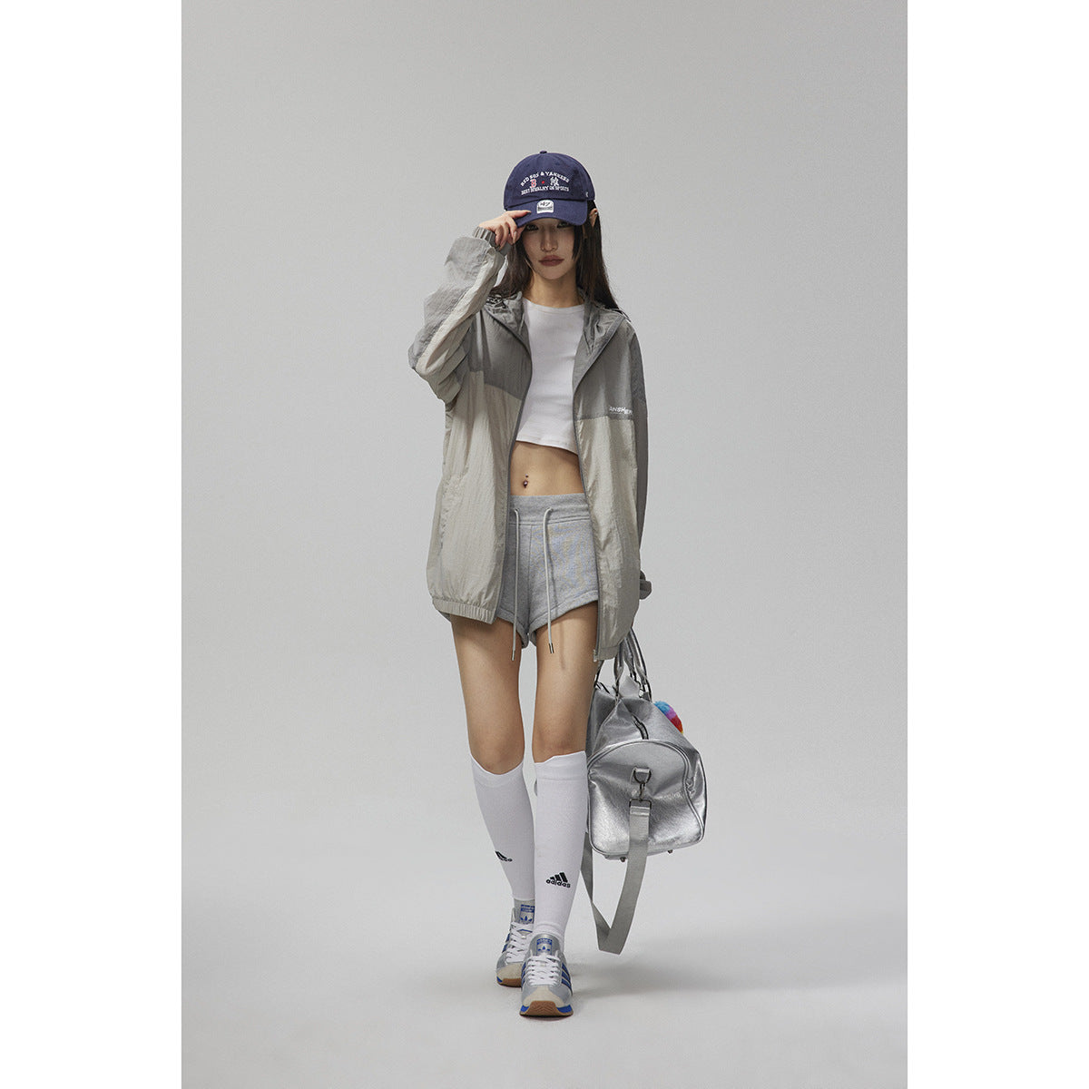 Model wearing a light gray hooded zip jacket with gray shorts, a navy cap, white socks and sneakers.