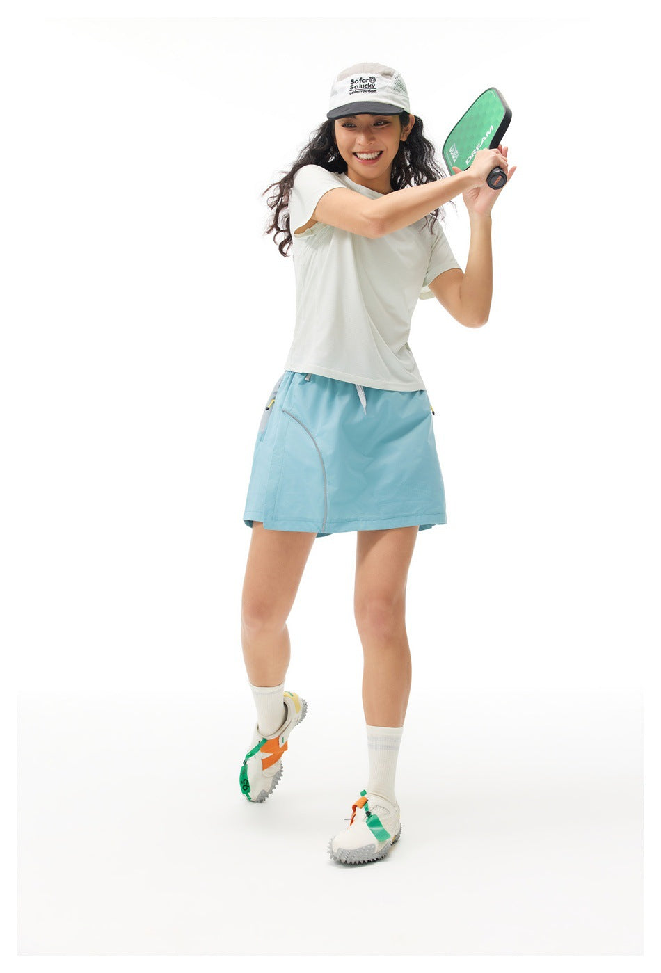 Model in an off-white short-sleeve shirt and light blue athletic skirt, swinging a paddle.