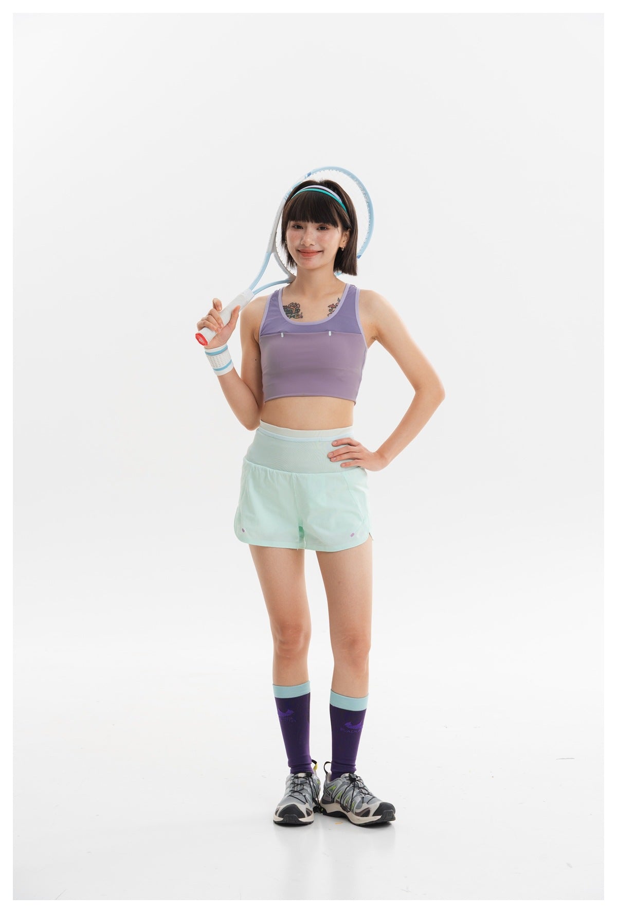 Woman in a lavender sports bra and mint shorts posing with a tennis racket on a white background.