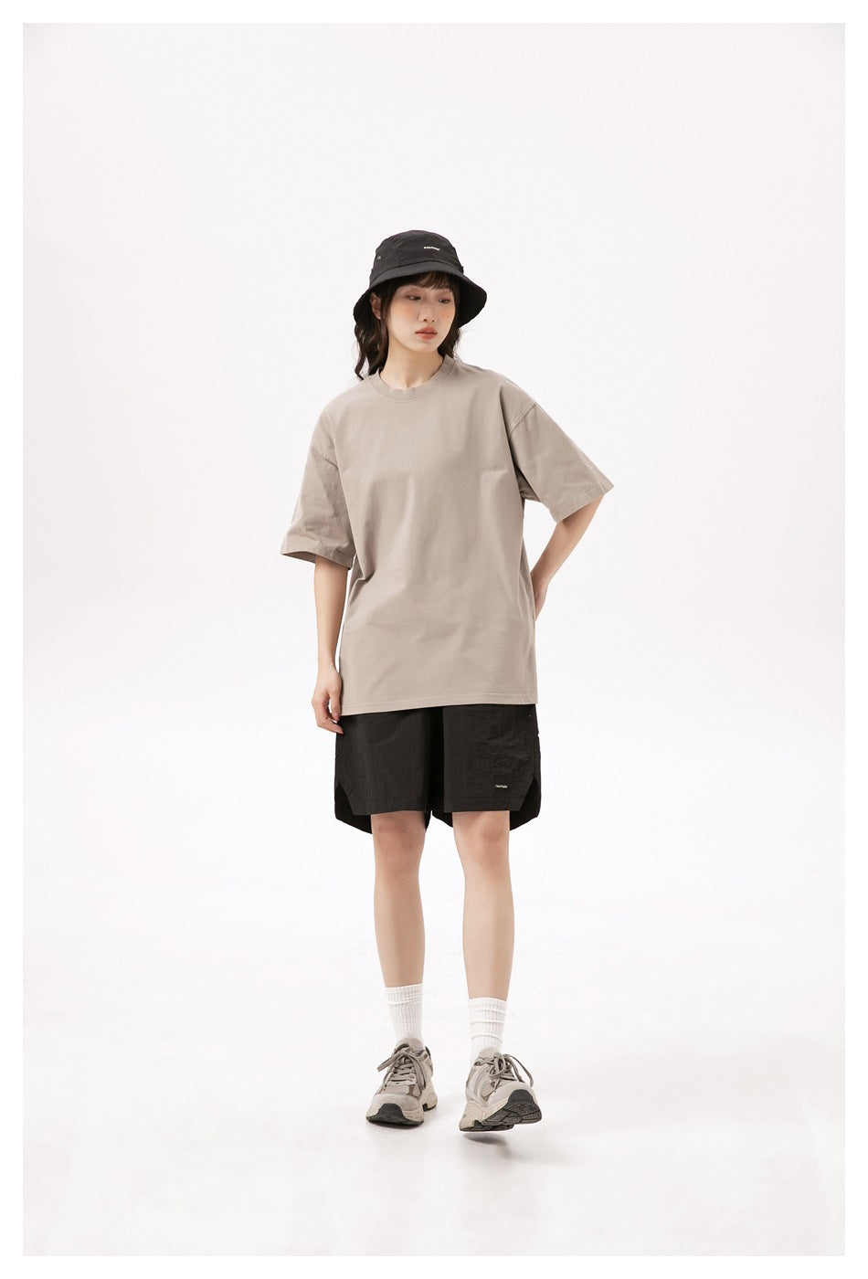 Model wearing a loose beige T-shirt, black shorts, and a black bucket hat, shown from the front.