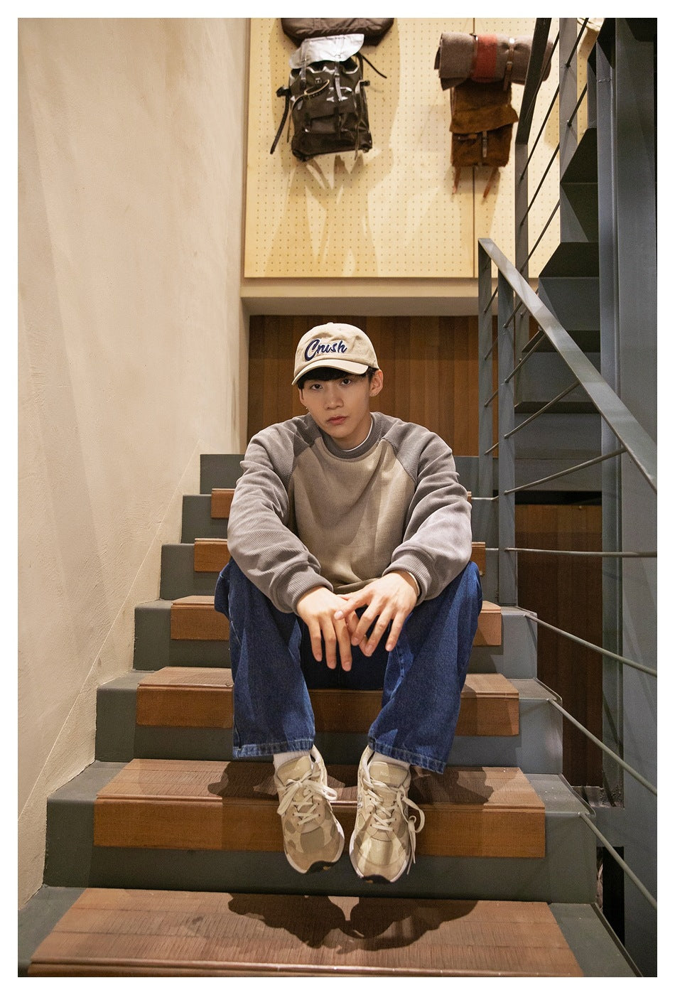 A seated man wears a taupe sweatshirt with gray raglan sleeves, dark blue jeans, and a beige embroidered cap.