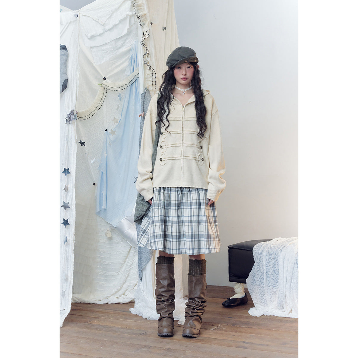 Model wearing a cream hooded zip sweater with loop-and-button detailing and a gray plaid skirt.