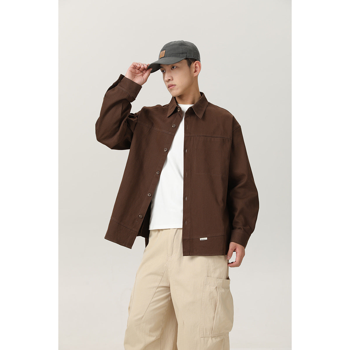 Model adjusts his cap while wearing an open dark brown shirt jacket over a white tee and cream cargo pants.