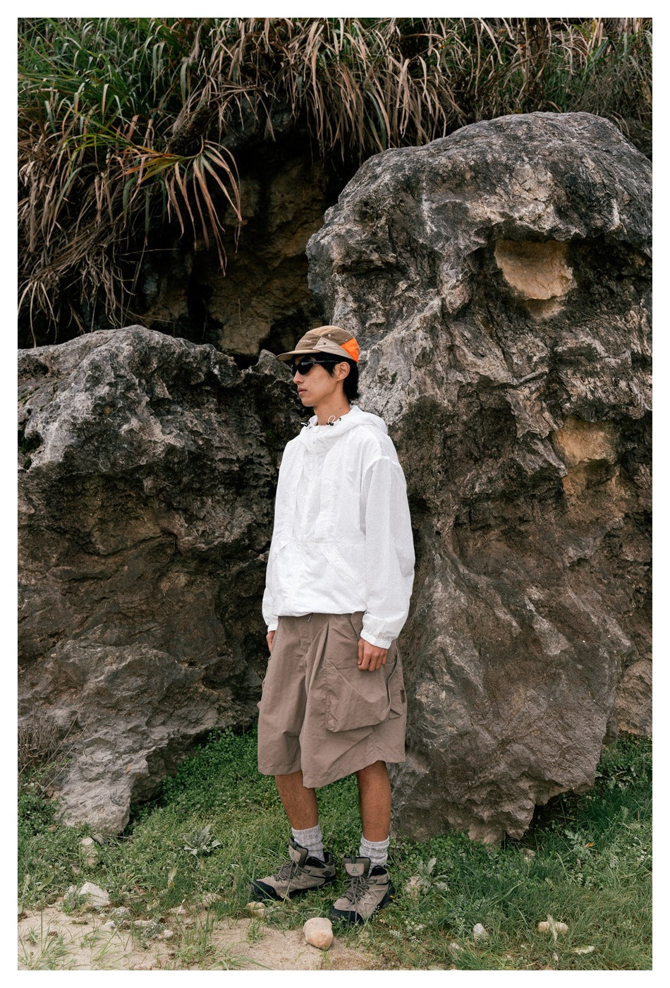 A man outdoors wears a white hooded pullover, taupe cargo shorts, hiking boots, sunglasses, and a color-block cap.