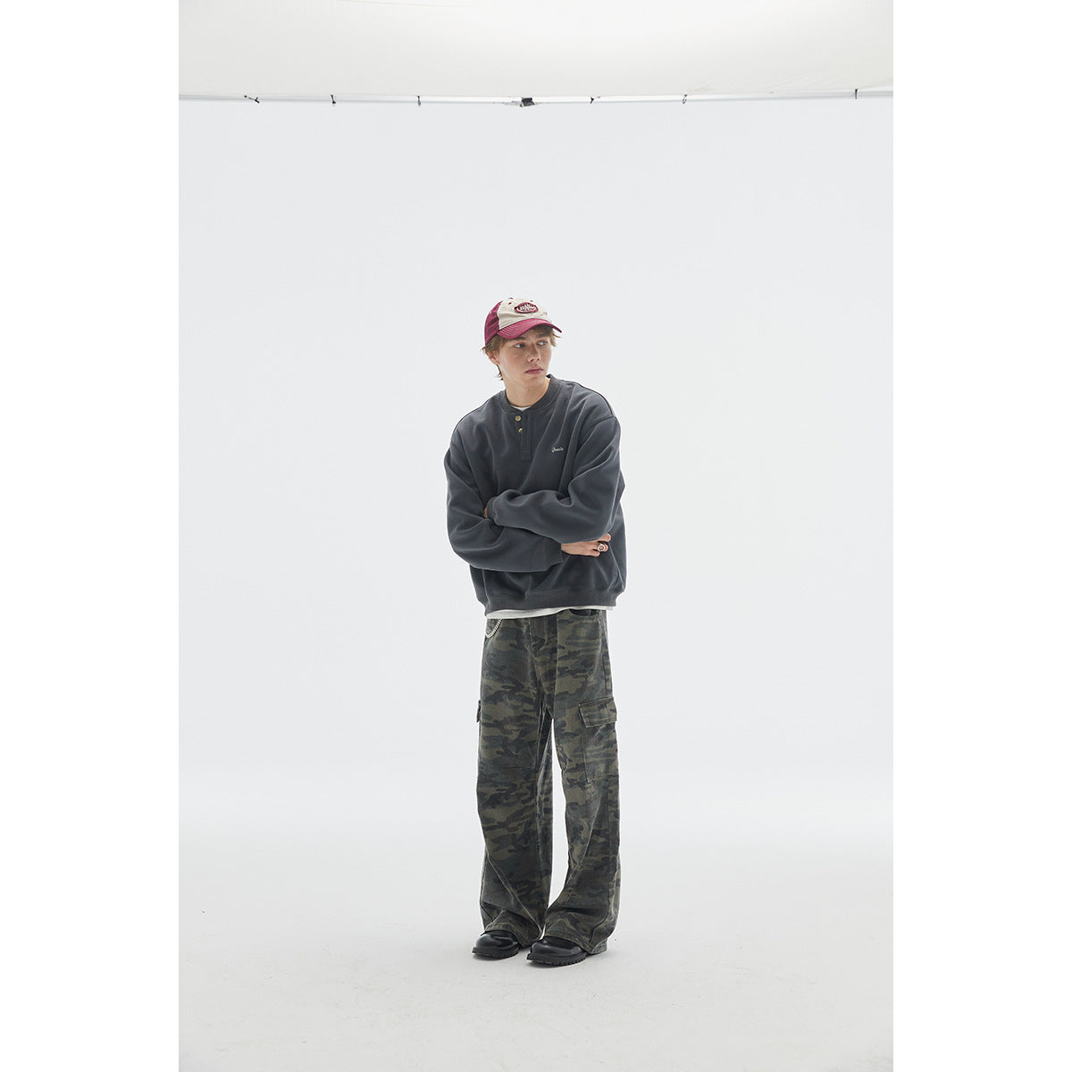Model wearing a charcoal henley sweatshirt with camouflage cargo pants and a red-and-white cap.