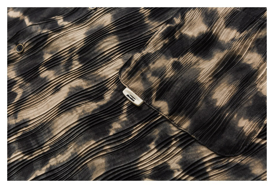 Close-up of a dark garment with beige abstract print, fine pleats, a button, and a small sewn label.