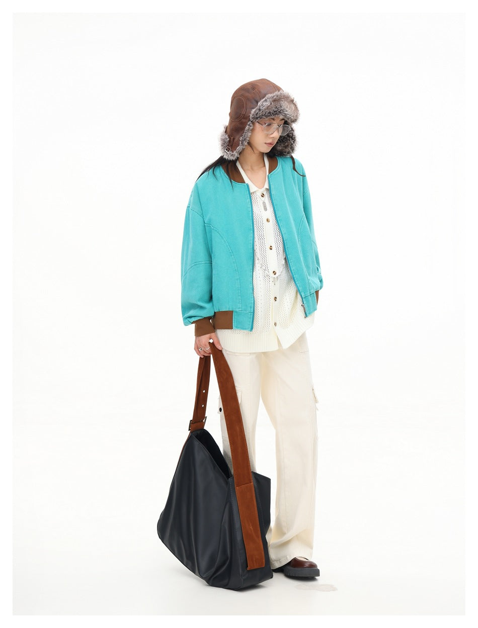 Model looking down, wearing teal bomber jacket with brown ribbed trim, cream pants and holding a large dark tote bag
