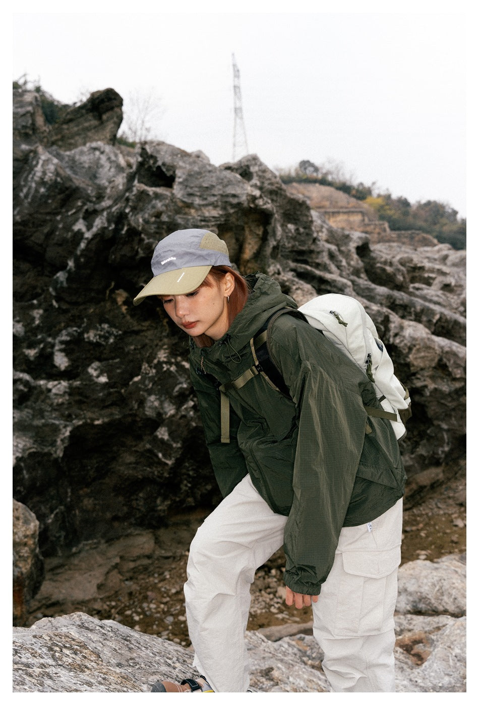 Close view of a model in a dark green hooded jacket and cream cargo pants, carrying a light-colored backpack.
