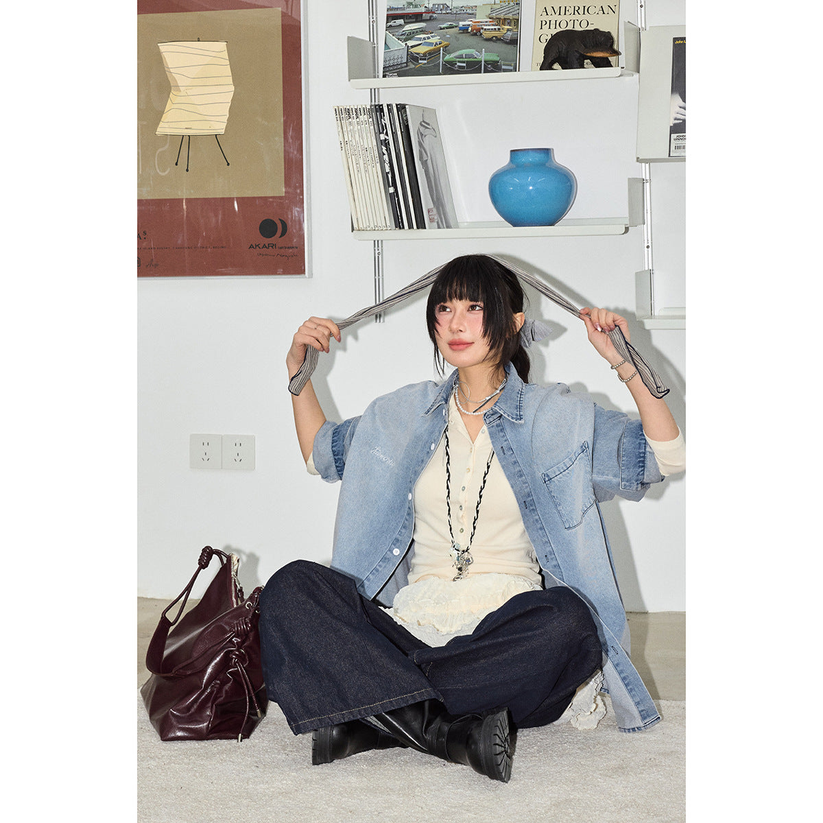Seated model in an oversized light blue denim shirt, cream layers and dark wide-leg jeans, holding the shirt sleeves outward.