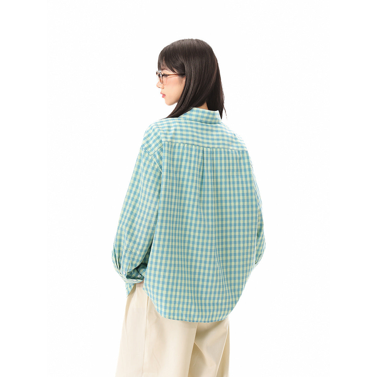 Rear view of a model wearing an oversized teal and pale yellow gingham shirt with a rounded hem and back pleat.