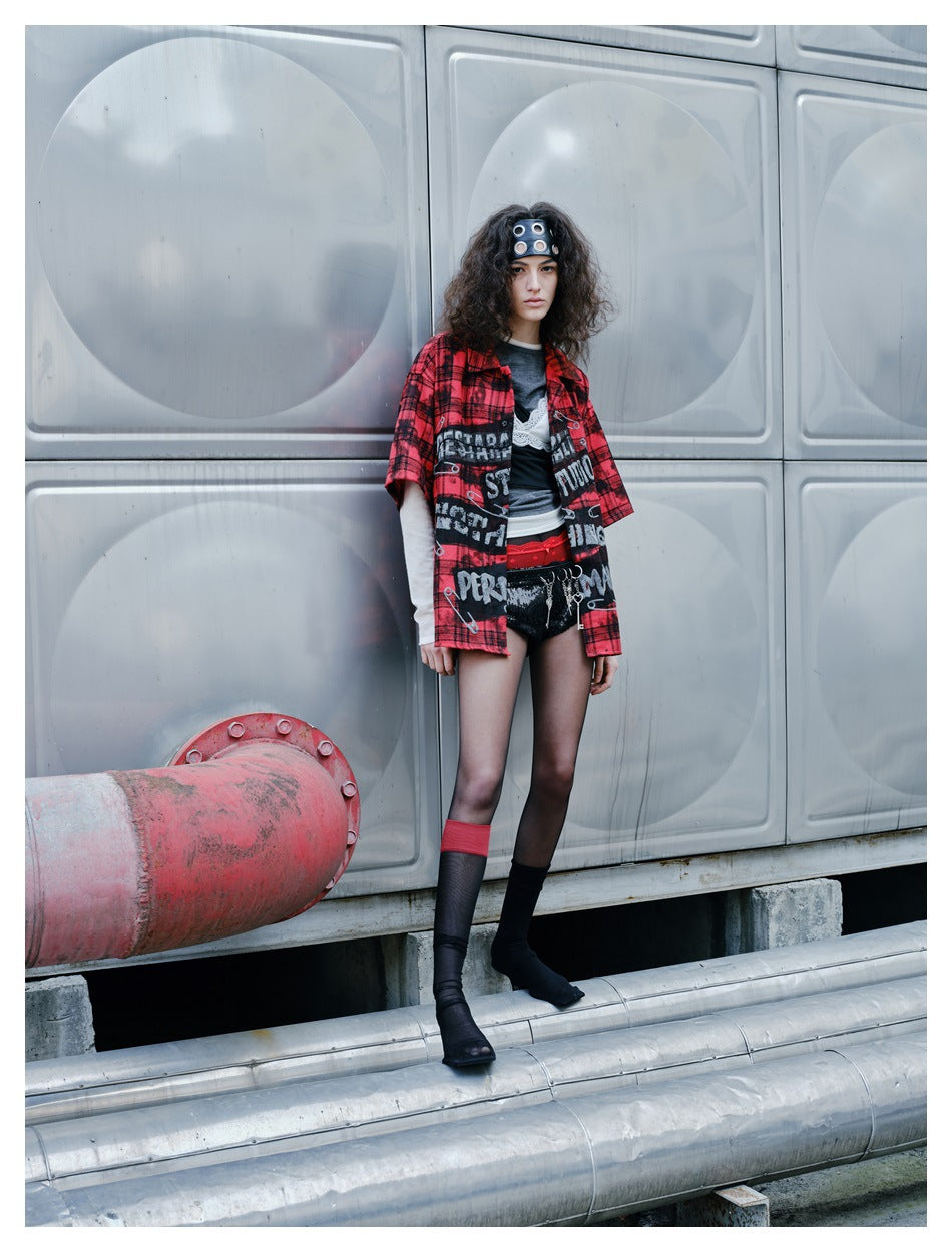 Full-length model wearing an oversized red-and-black plaid graphic shirt, black shorts, and dark knee-high socks.