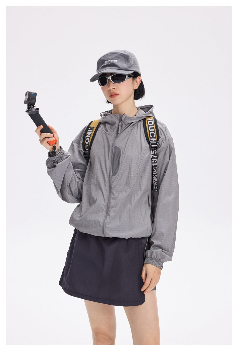Model in a gray hooded jacket and dark wrap skirt, shown from the front with a cap and sunglasses.