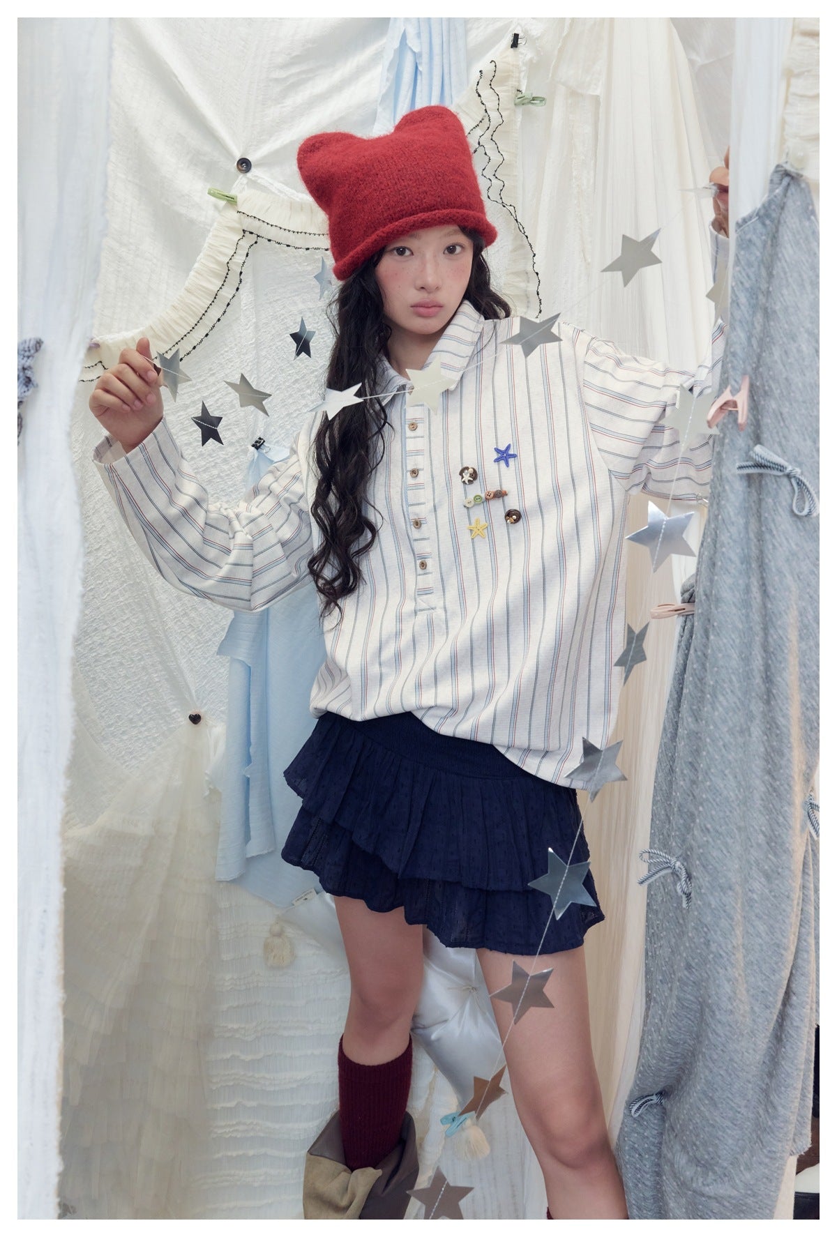 Model wearing the striped collared shirt over a navy tiered mini skirt with a red knit hat, among star garlands