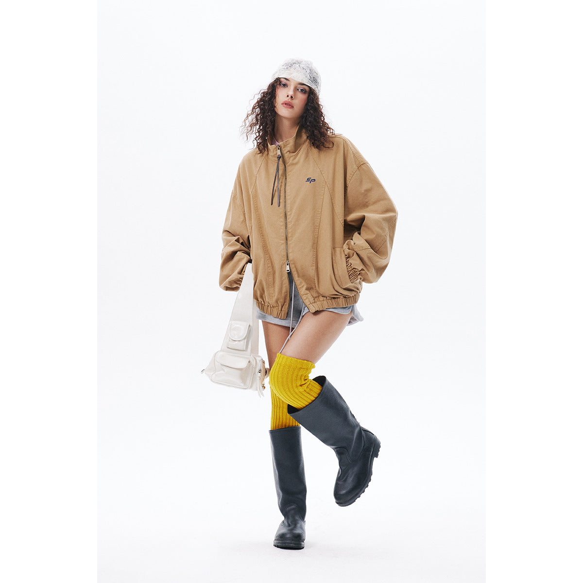 Model wearing an oversized tan zip jacket with elastic cuffs and hem, gray shorts, yellow socks and black boots.