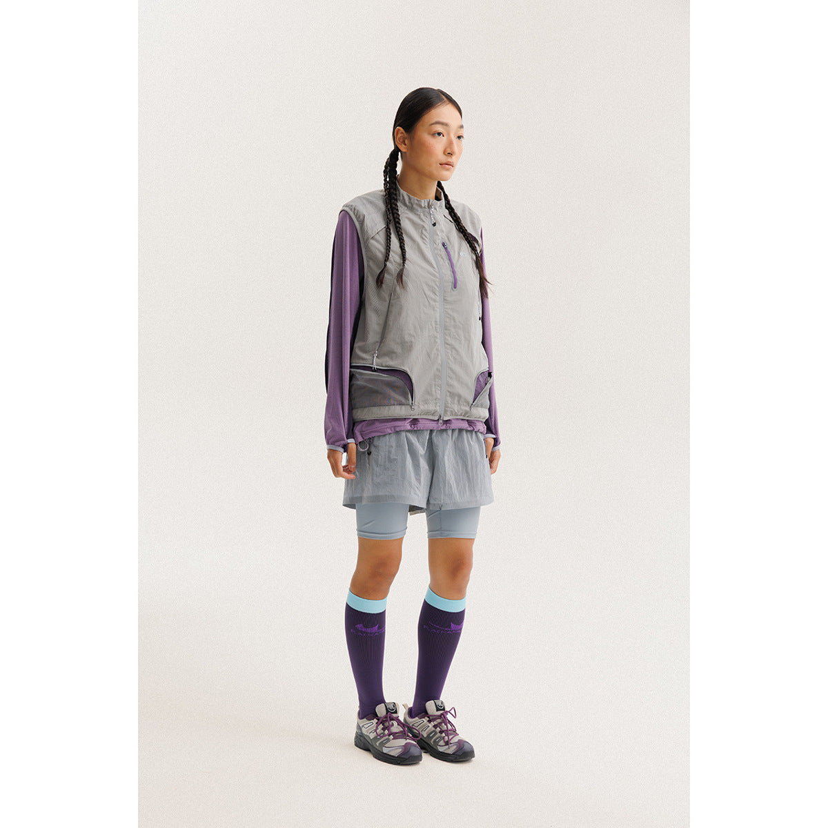 Model wearing a light gray zip-front utility vest over a purple long-sleeve top and gray athletic shorts.