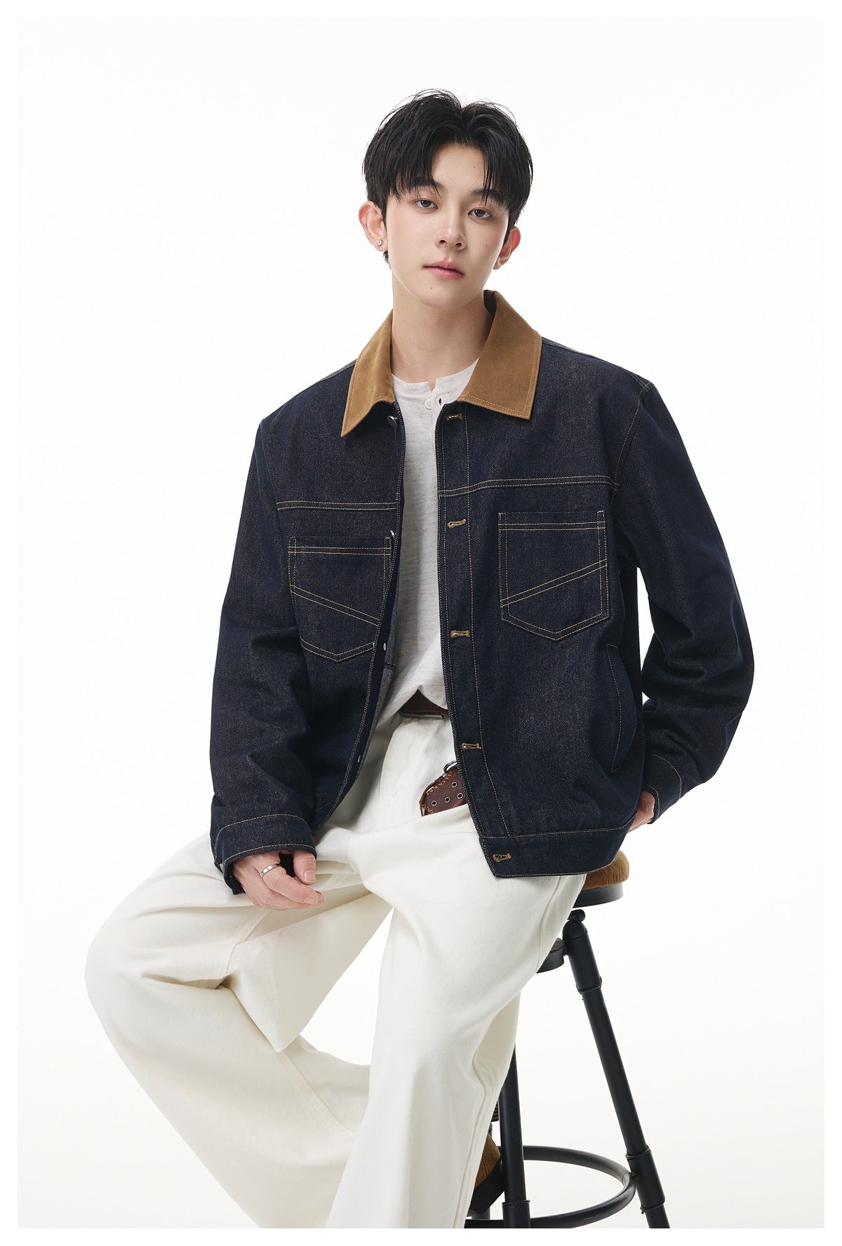 Model seated on a stool wearing a dark denim jacket with brown collar, white henley and cream pants