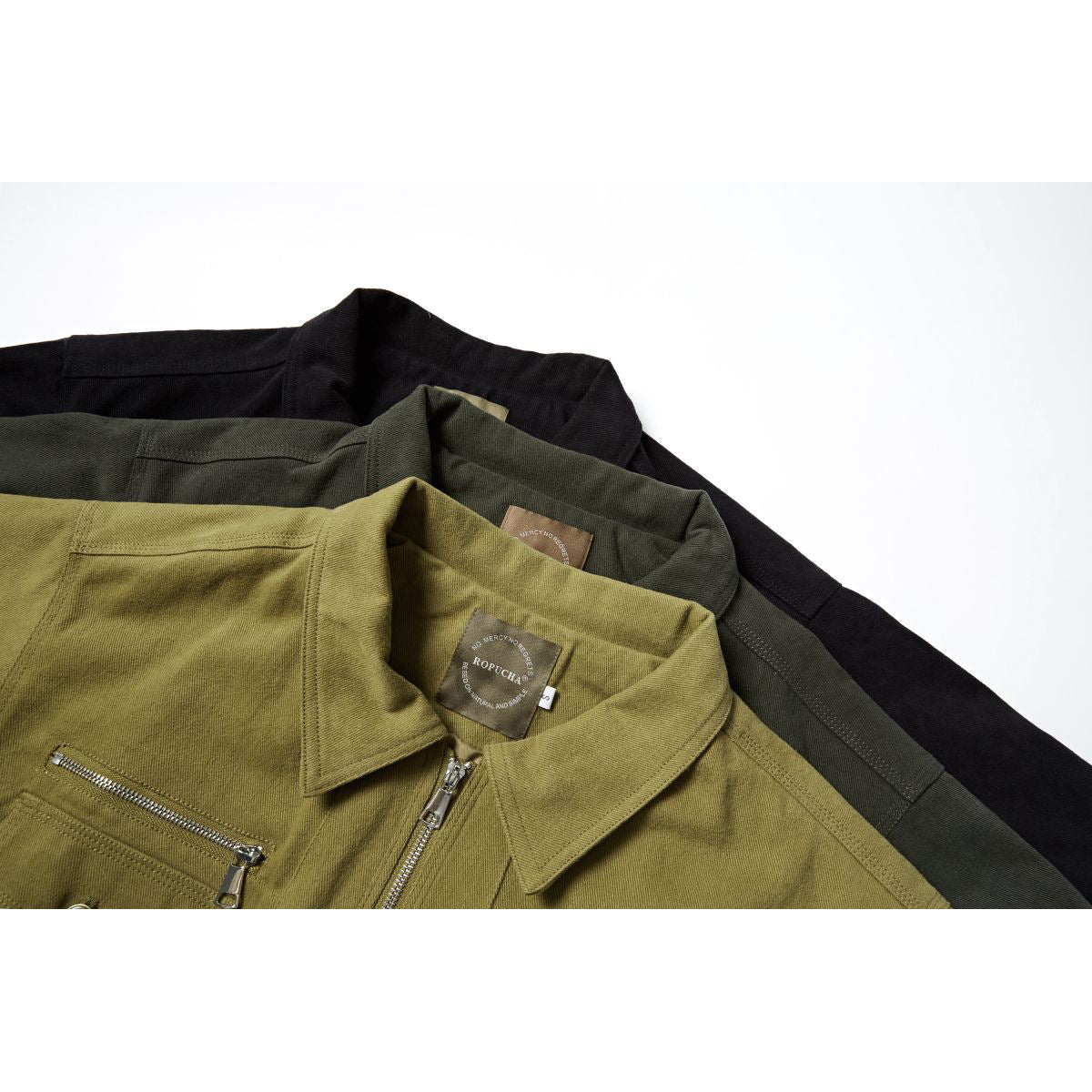 Close-up of three work jackets in black, dark olive, and mustard, showing collars, neck labels, and metal zippers.