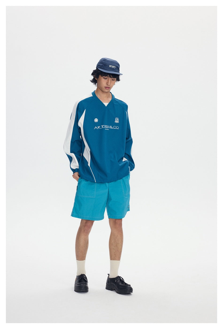 Model wearing blue and white color-block V-neck pullover with turquoise shorts, navy cap and black shoes, front view