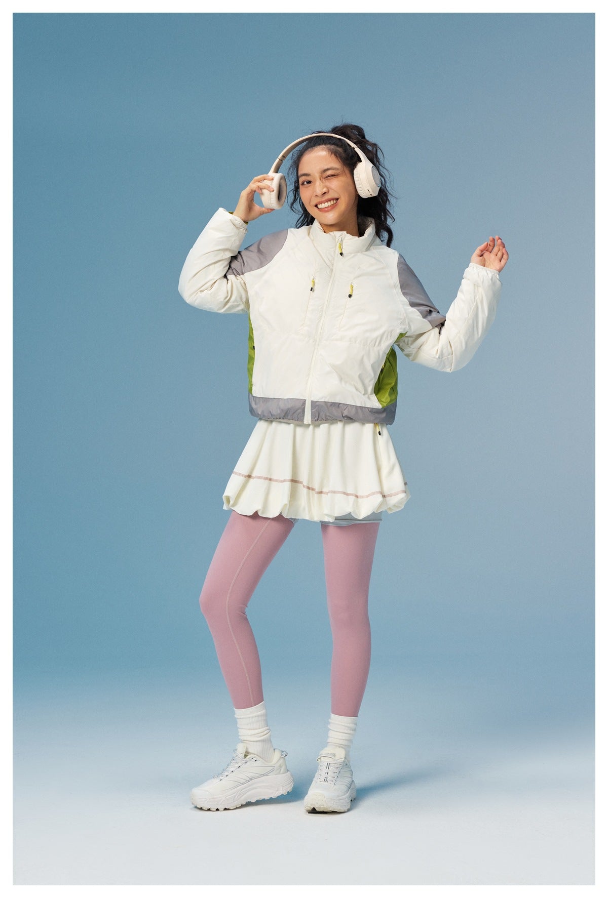 Full-length view of a woman wearing a white jacket with gray panels and lime accents, a white skirt, and pink leggings.