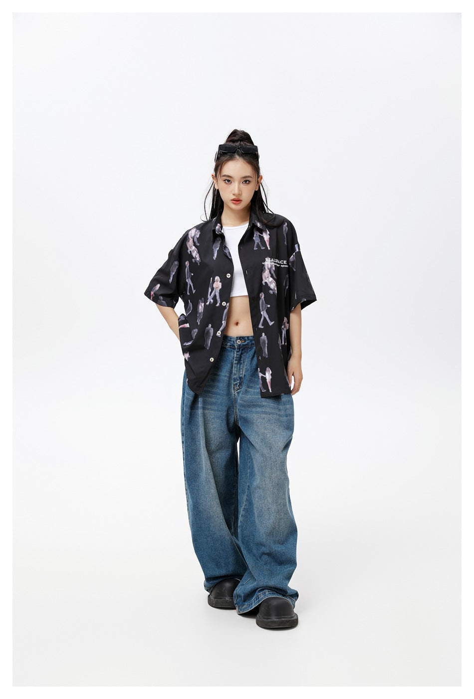 Model wearing black printed short-sleeve shirt open over a white crop top with wide-leg blue jeans