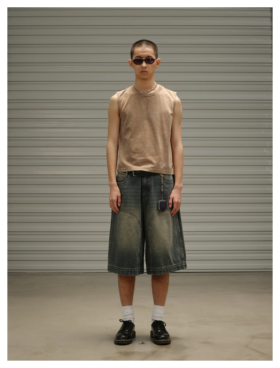Front view of a man wearing a tan sleeveless top and loose, faded dark denim shorts.
