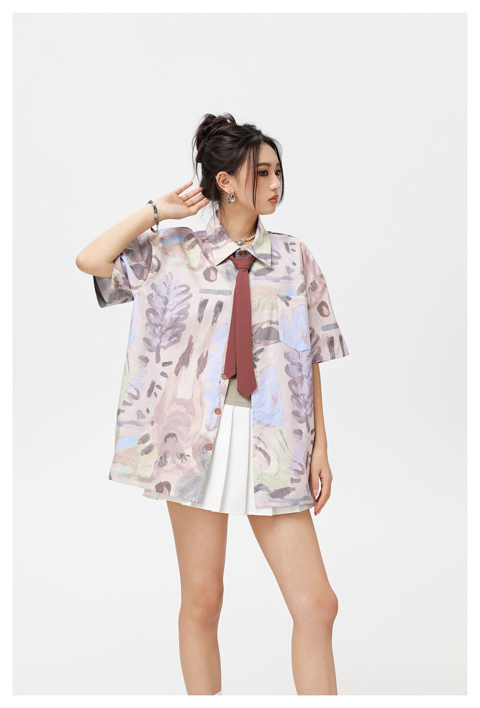 Model in a pastel abstract-print oversized short-sleeve shirt with chest pocket, burgundy tie and white pleated skirt.
