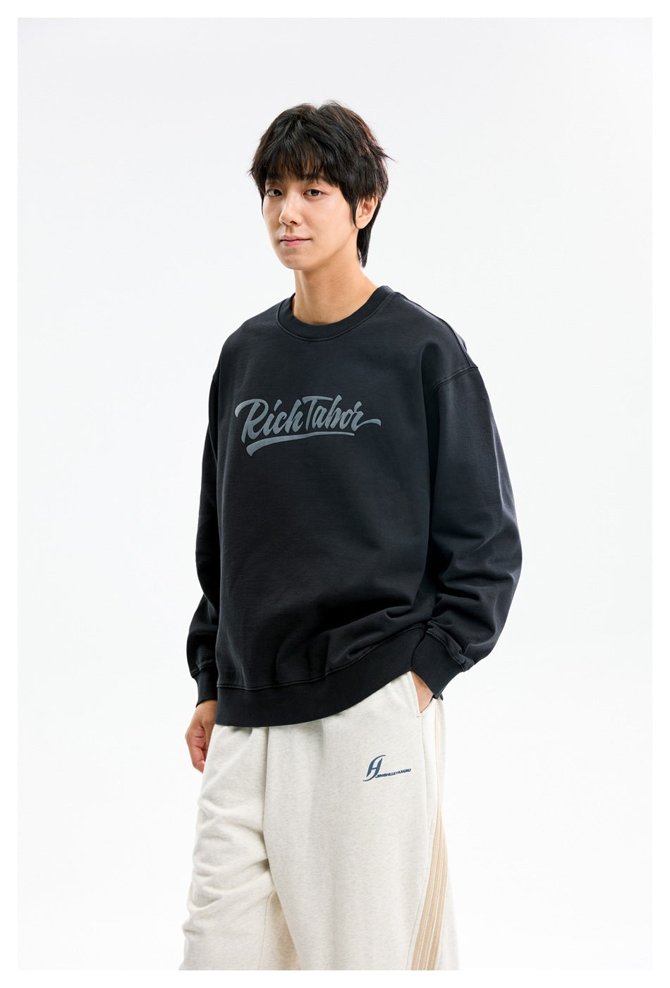 Model wearing a black crewneck sweatshirt with gray script chest print and light sweatpants.