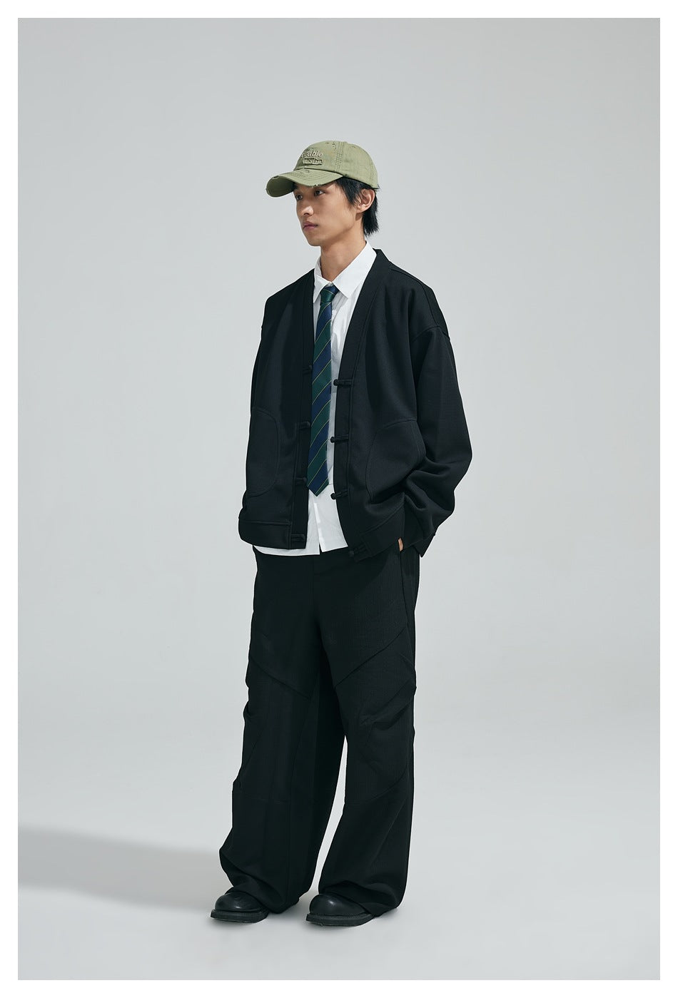 Model wearing black frog-button cardigan jacket over white shirt and tie with wide black pants and green cap, full length