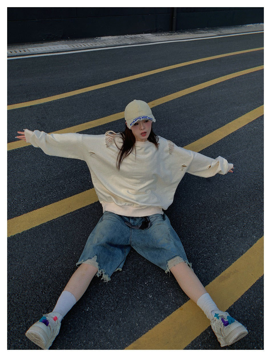 Model sitting on a road with arms outstretched in a khaki distressed sweatshirt and denim shorts