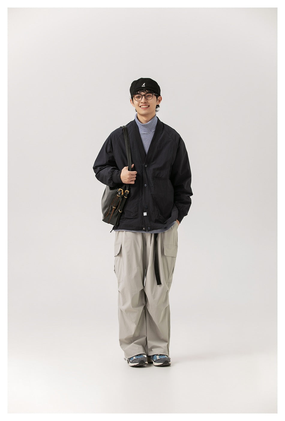 Full-length view of model in a black V-neck snap-front jacket, striped turtleneck, gray cargo pants and a shoulder bag