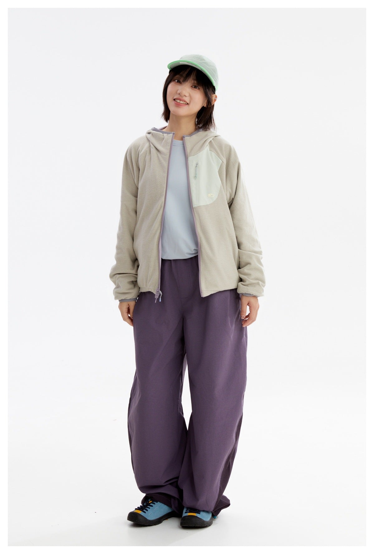 Model wearing a light gray-beige hooded zip jacket with a pale mint chest pocket and dark purple trousers.