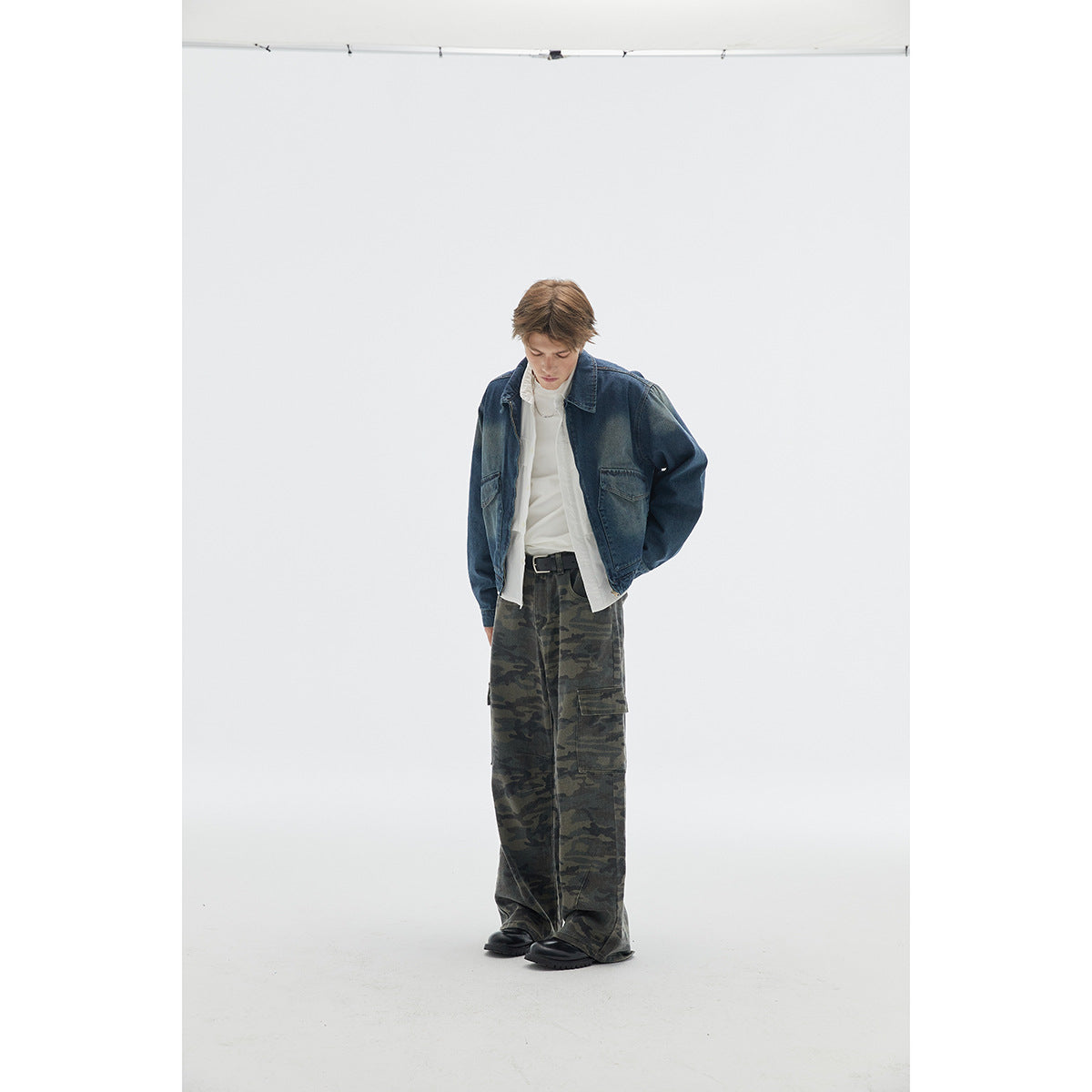 Model wearing a dark blue denim jacket over white layers and loose camouflage cargo pants, looking down.