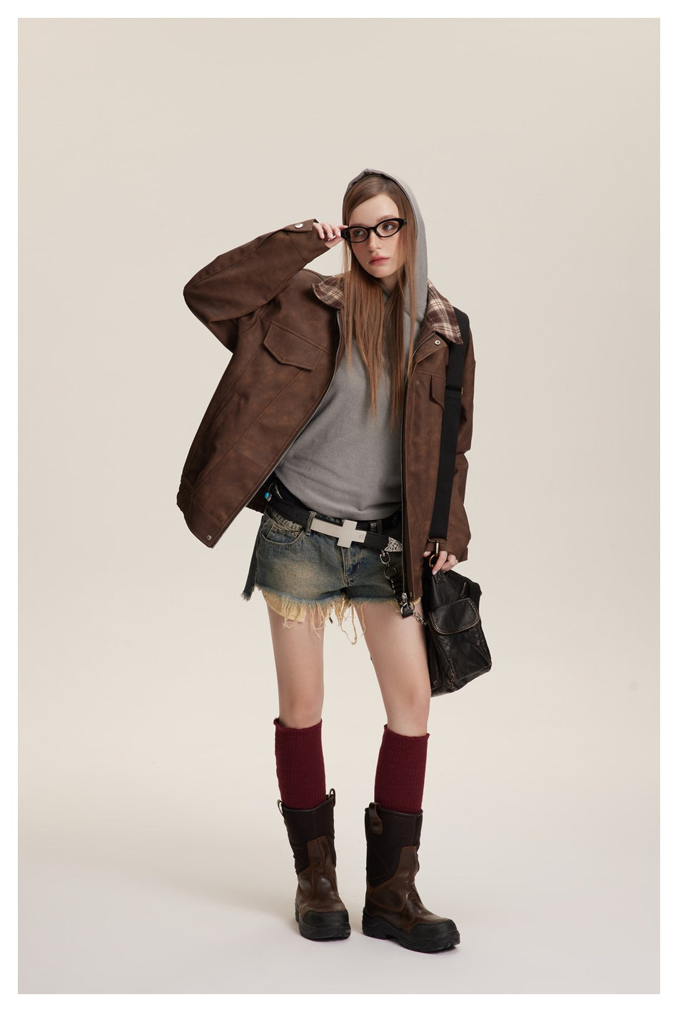Model wearing the brown jacket open over a gray hoodie with denim shorts, red knee socks and boots, hand raised to glasses