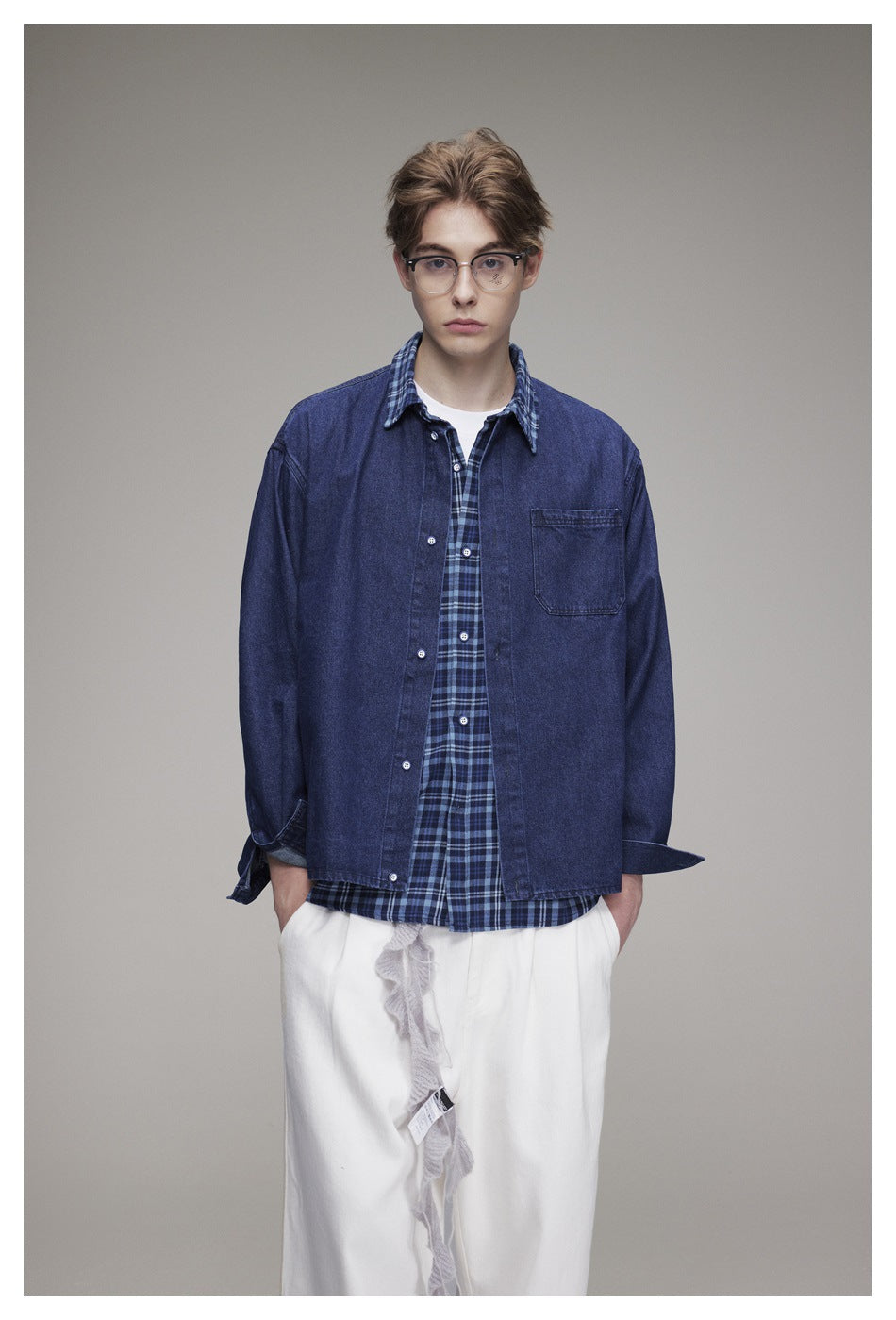 Front view of a model in a navy denim overshirt with a chest pocket, layered over a blue plaid shirt and white trousers.