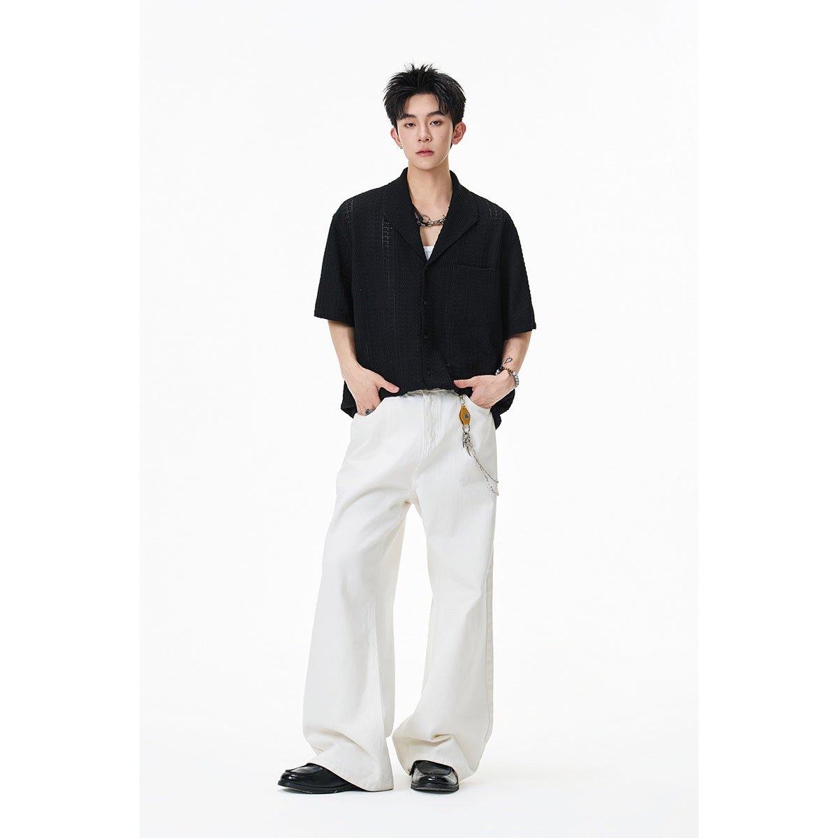 Model wearing a black short-sleeve textured button-up shirt with a chest pocket and wide white trousers.