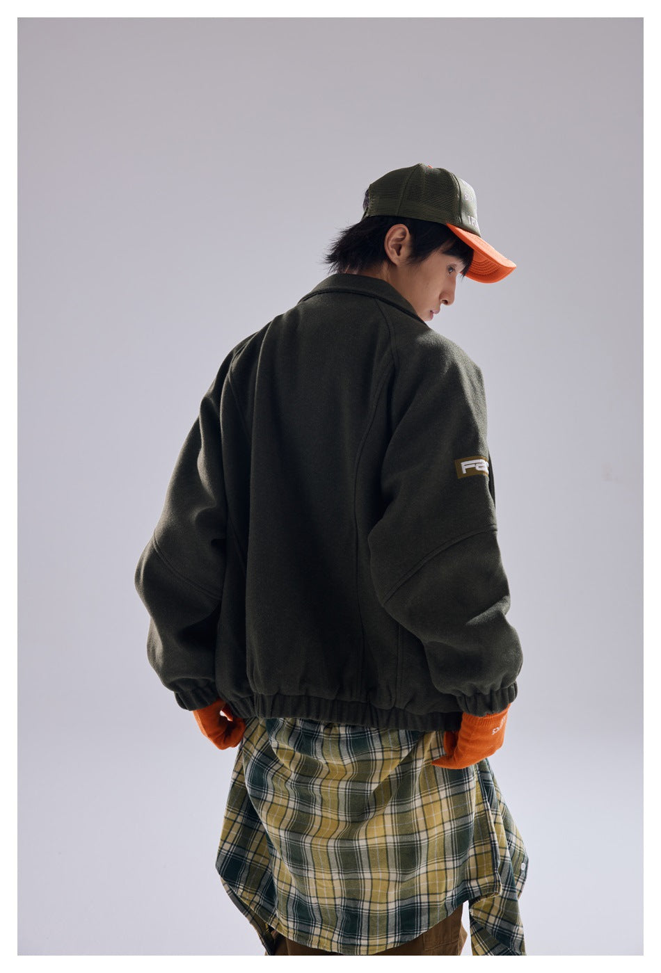 Rear view of a model wearing a dark olive jacket over a long green, cream, and yellow plaid shirt.